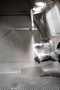 A person wearing protective gloves uses a sandblasting machine inside a ventilated enclosure to clean or finish a metal object, with dust and light visible in the workspace.