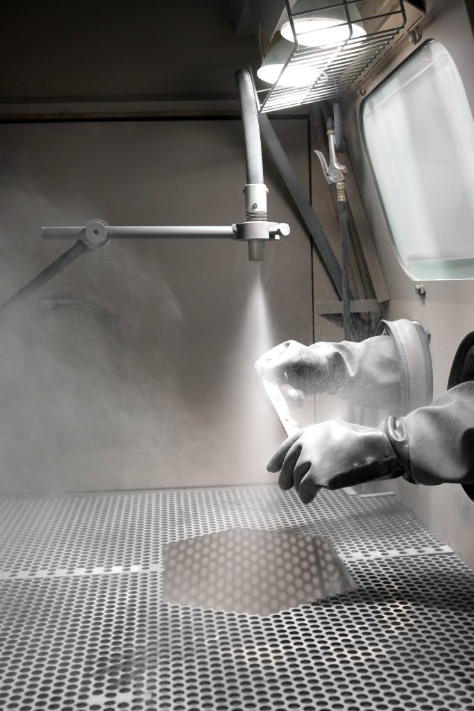 A person wearing protective gloves uses a sandblasting machine inside a ventilated enclosure to clean or finish a metal object, with dust and light visible in the workspace.