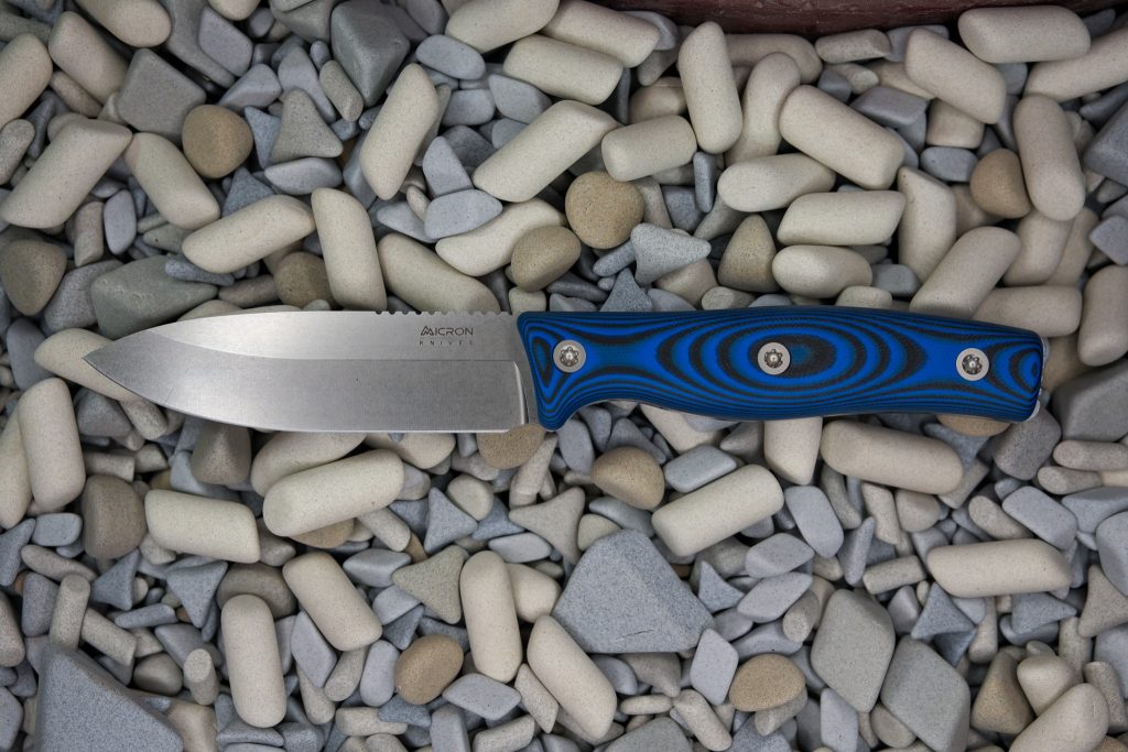 A fixed-blade knife with a blue and black patterned handle lies on a bed of smooth, assorted gray and white pebbles and stones.