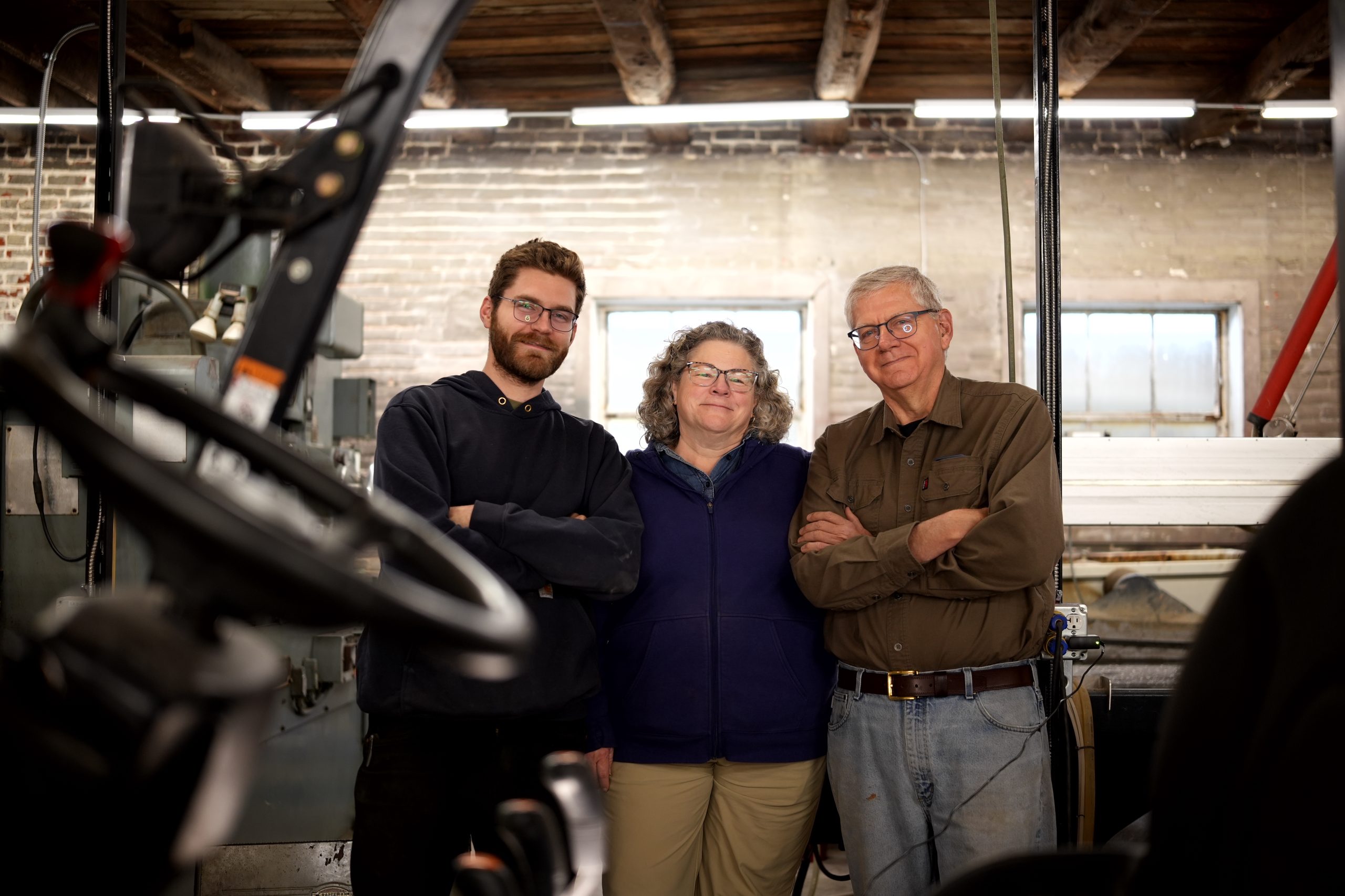 Three people—two men and one woman—stand side by side in a workshop, smiling with their arms crossed. Exposed wooden beams and machinery in the background tell a story about their creative workspace.