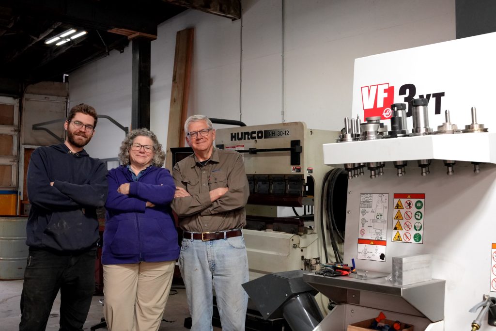 Three people stand smiling with arms crossed in a workshop, surrounded by industrial machinery and equipment, including a white machine labeled VF 3YT. The atmosphere is friendly and collaborative.