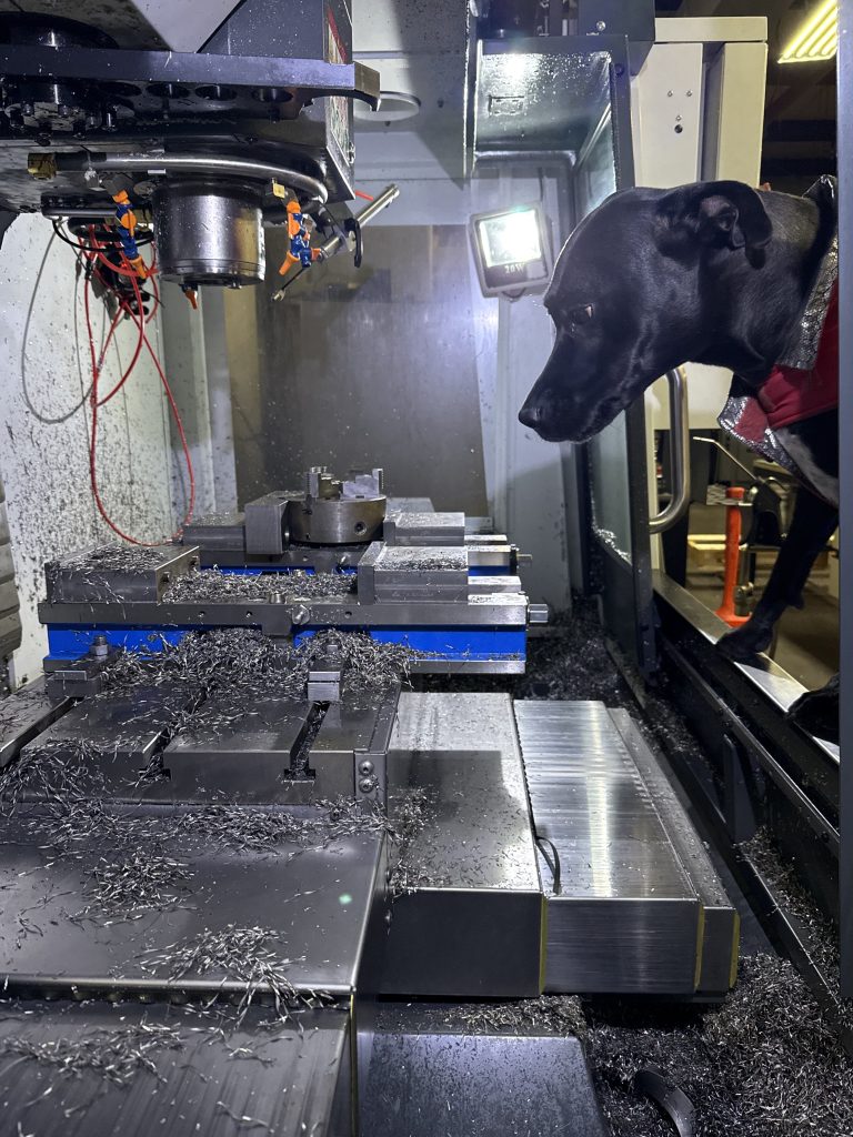A black dog curiously looks at a CNC milling machine in operation, with metal shavings scattered about the work surface and equipment parts visible under bright lighting.