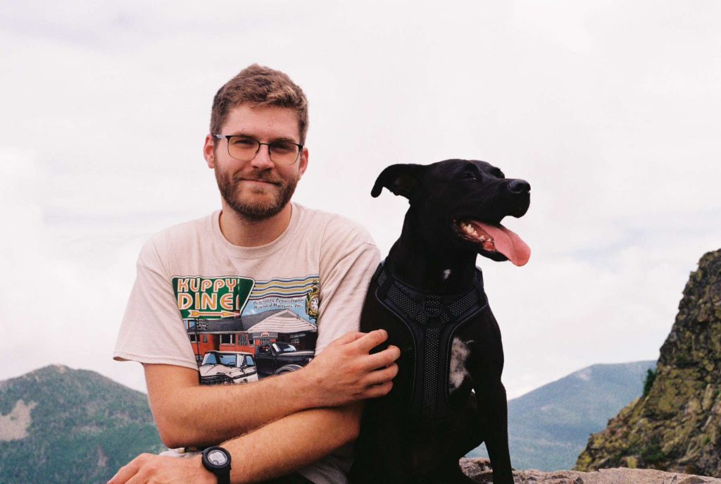 A man with glasses and a beard, wearing a Huppy Diner T-shirt, sits on a rocky ledge beside his black dog with its tongue out. Mountains rise behind them under a cloudy sky—an adventure that comes with no warranty.