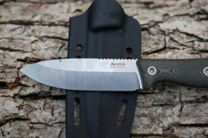 A Micron MK-4 fixed-blade knife with a textured black handle rests on its black sheath, set atop rough tree bark.