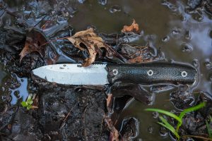 A fixed-blade knife with a dark handle lies on wet, muddy ground surrounded by fallen leaves and small plants. About half of the blade is partially covered with water droplets and mud.