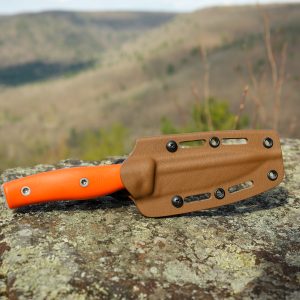 The Micron MK-4, a fixed-blade knife with an orange handle and brown sheath, rests on a large rock against a blurred backdrop of hilly forest.