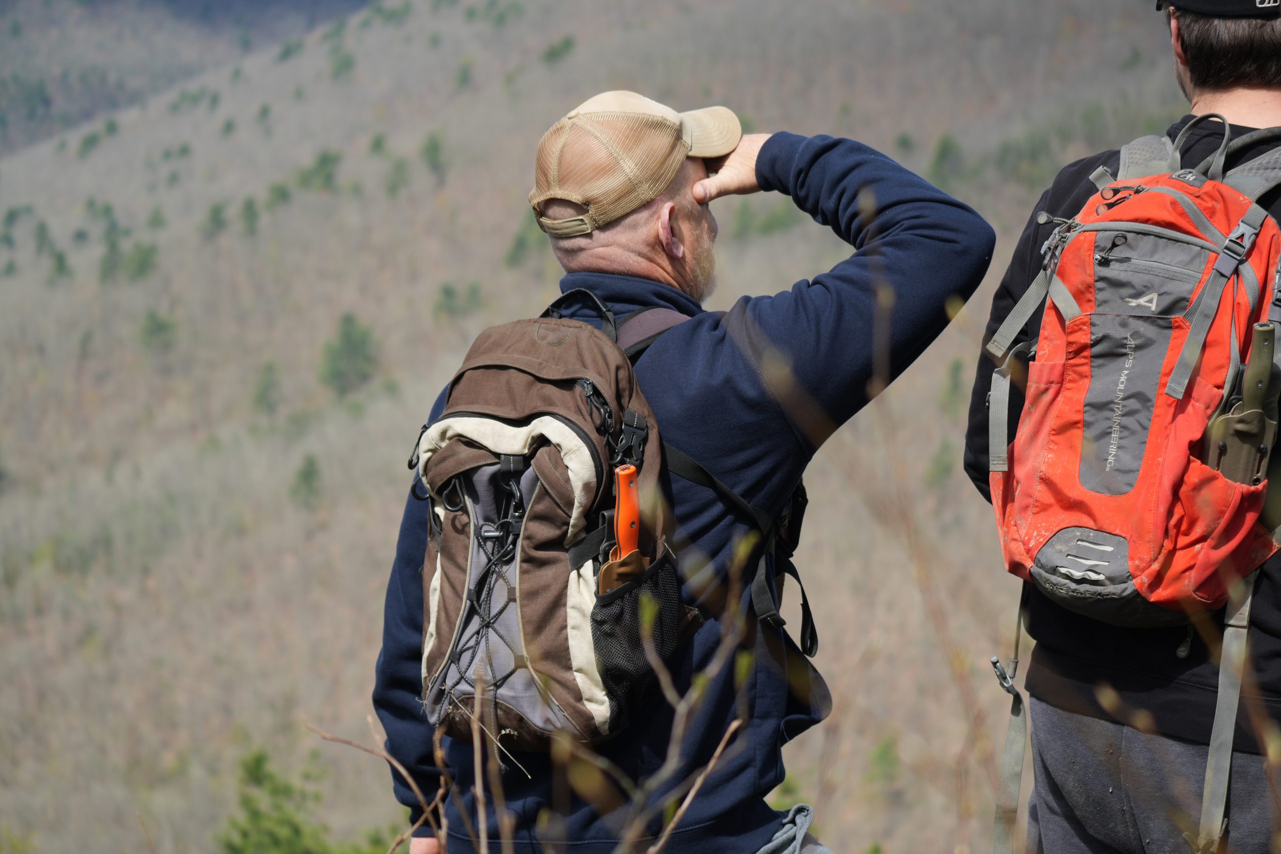 Two hikers with backpacks stand on a ledge, one shielding his eyes and looking out at a forested valley below. The scene suggests exploration and adventure in the mountains.