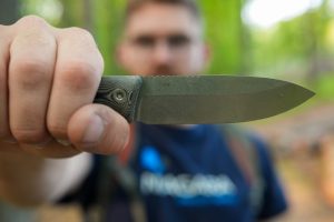 A person holds a Micron MK-4 knife toward the camera, its blade sharply in focus while their face is blurred in the forested outdoor background.