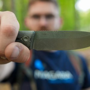 A person holds a Micron MK-4 knife toward the camera, its blade sharply in focus while their face is blurred in the forested outdoor background.