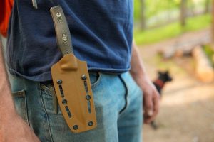 A person wearing blue jeans and a navy t-shirt has a large knife in a tan sheath attached to their belt. The background is outdoors, slightly blurred, with a black dog visible in the distance.