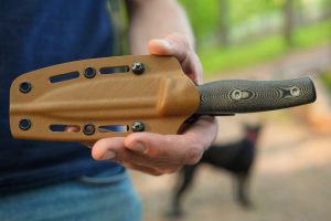 A person holding a sheathed Micron MK-4 knife with a textured handle outdoors, surrounded by greenery, with a black dog visible in the distance.