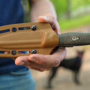 A person holding a sheathed Micron MK-4 knife with a textured handle outdoors, surrounded by greenery, with a black dog visible in the distance.