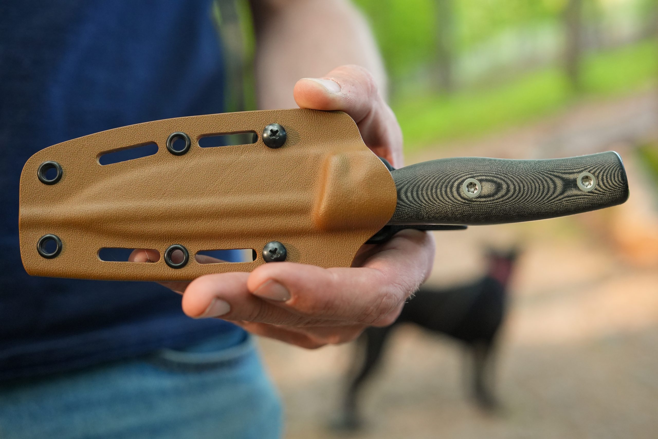 A person holding a sheathed Micron MK-4 knife with a textured handle outdoors, surrounded by greenery, with a black dog visible in the distance.