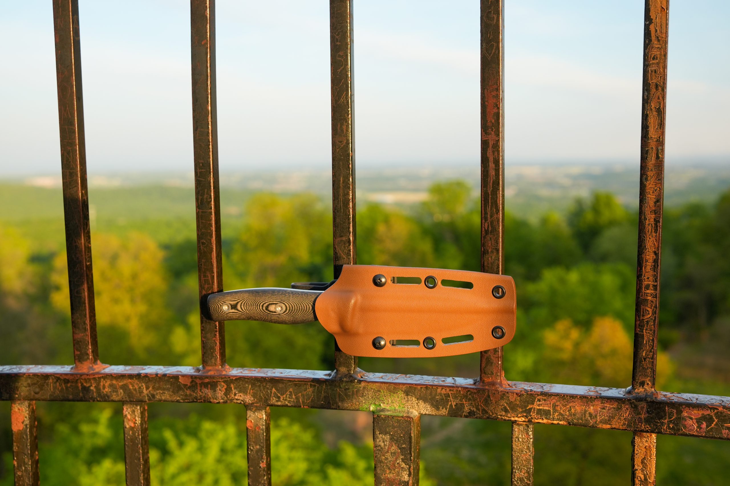 A fixed-blade knife in a brown sheath is wedged horizontally between the vertical bars of a rusty metal fence, with a scenic, green forest landscape visible in the blurry background.