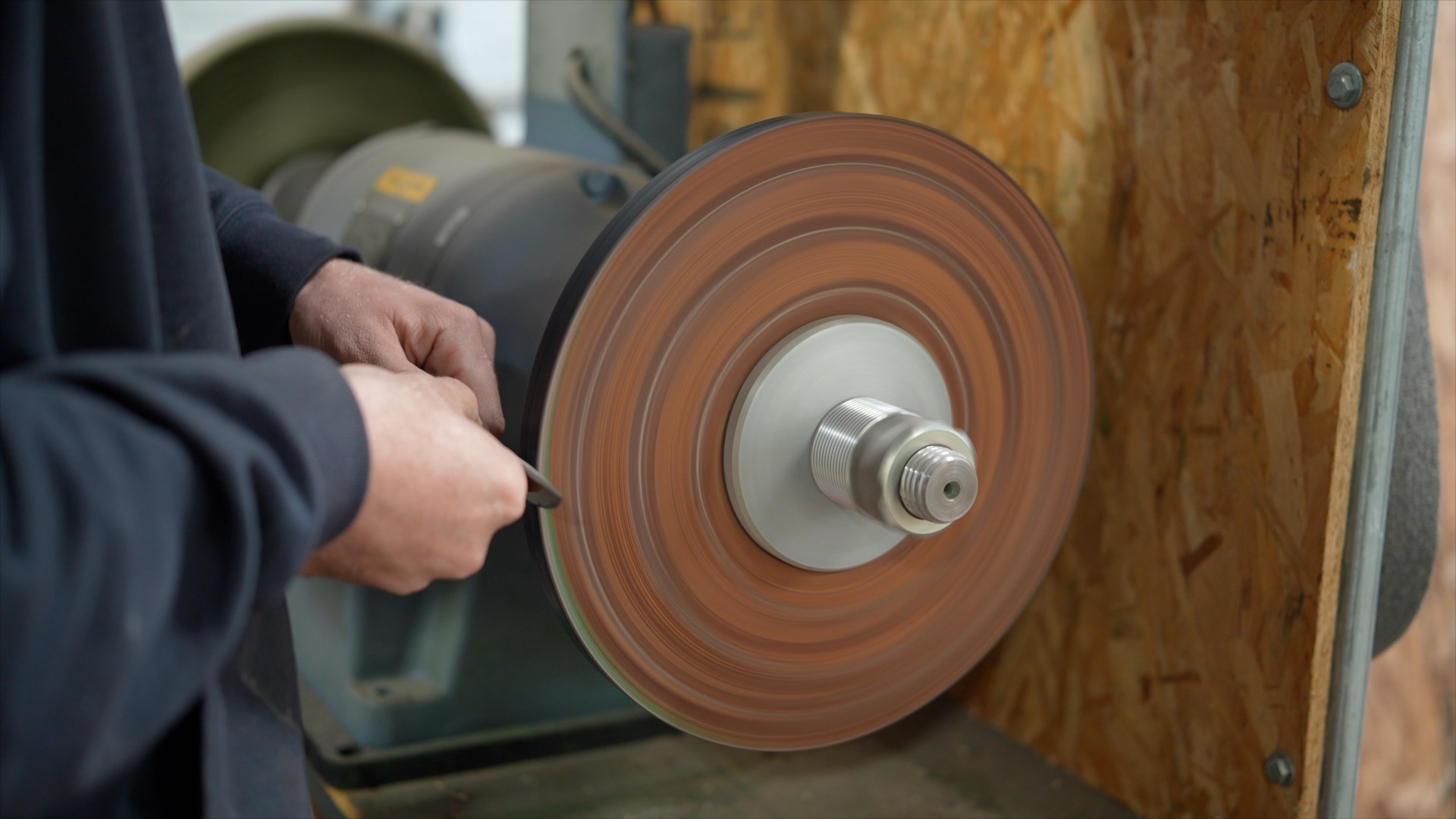 A person sharpens a tool on a rotating grinding wheel, with their hands close to the spinning abrasive surface, inside a workshop setting where proper use helps maintain warranty coverage.