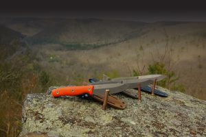 Two large knives with sheaths, one featuring an orange handle and backed by a warranty, rest on a rock ledge overlooking a vast, hilly landscape with dry vegetation and distant hills under a cloudy sky.