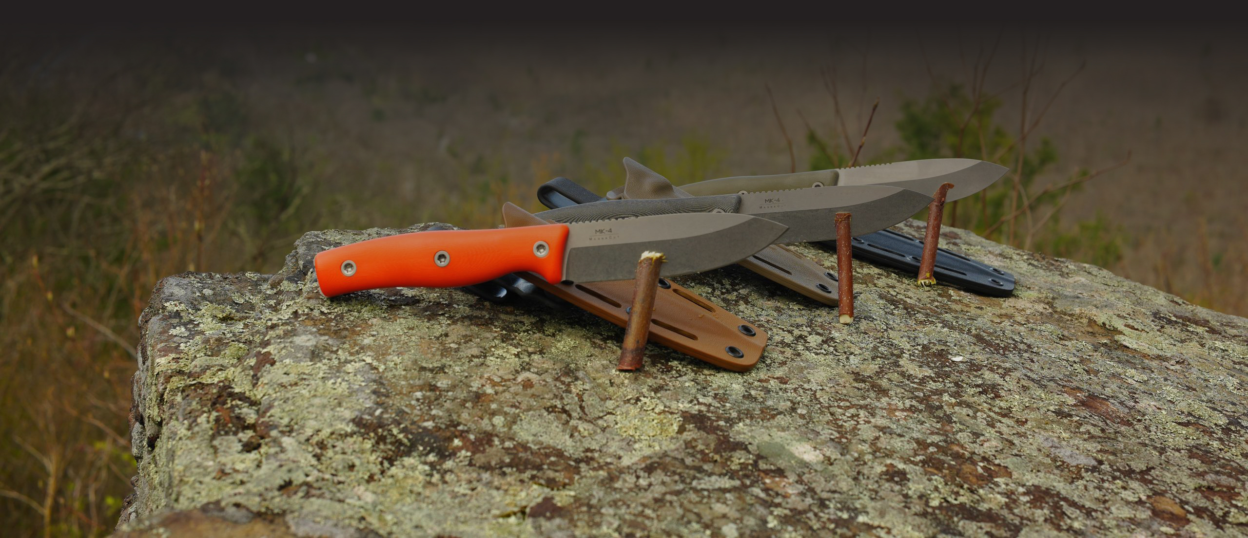 Three fixed-blade knives with colored handles and sheaths, backed by a product warranty, are displayed outdoors on a large, weathered rock with rusty nails standing upright nearby and blurred natural scenery in the background.